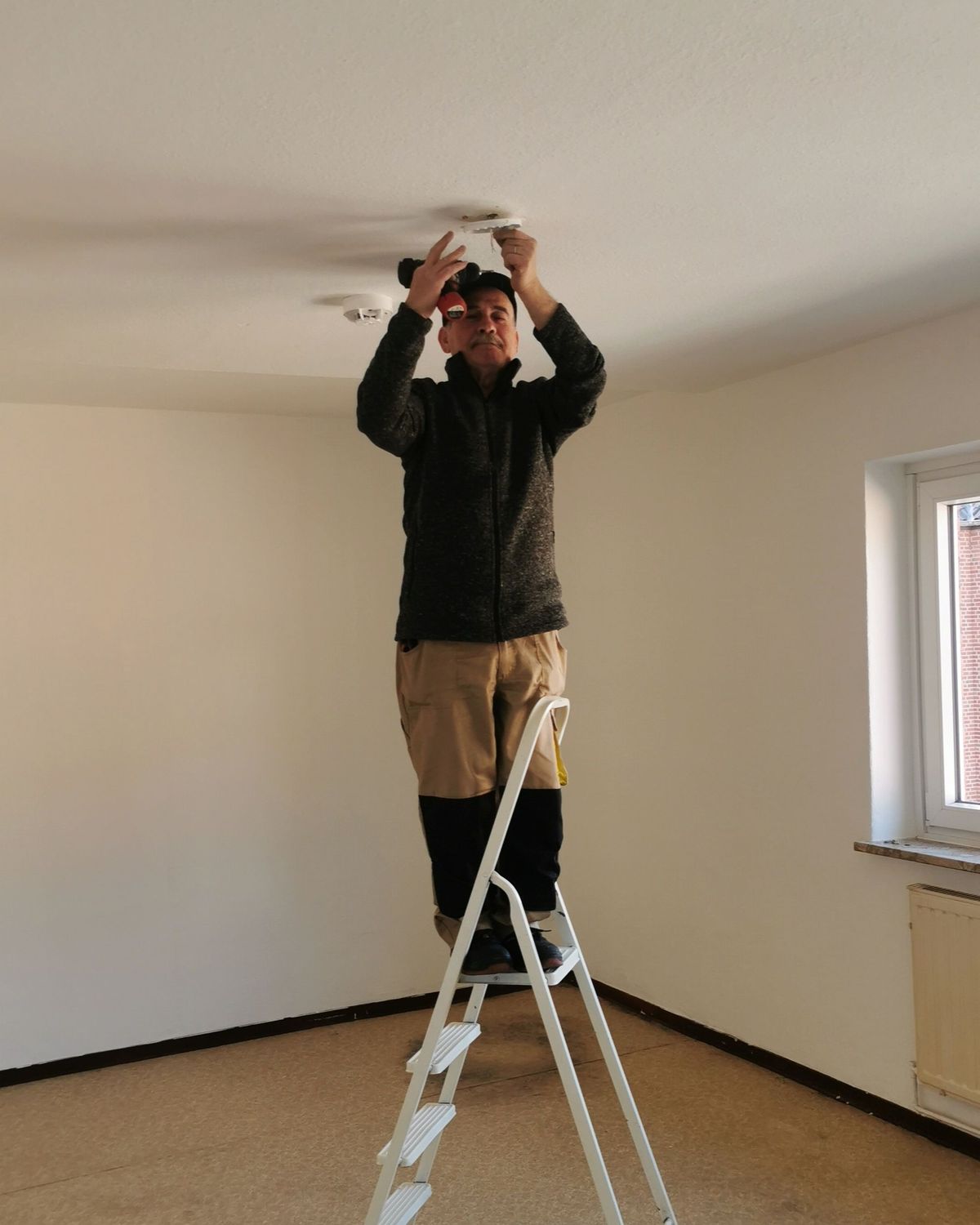 Een man op een trapladder sluit een plafondlamp aan in een lege kamer.